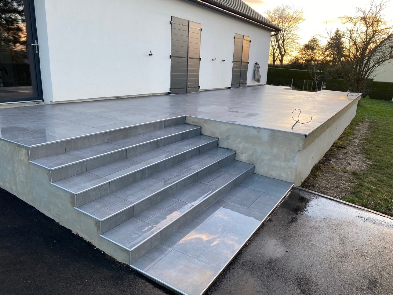 Une grande terrasse extérieure en carrelage gris avec des marches, récemment terminée. La terrasse est attachée à une maison blanche avec des volets gris.
