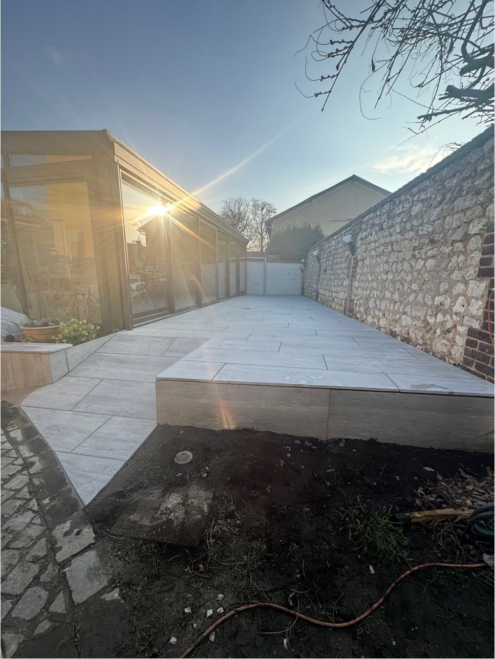 Une terrasse en carrelage gris clair avec un mur en pierre à l'arrière-plan, baignée par la lumière du soleil couchant.