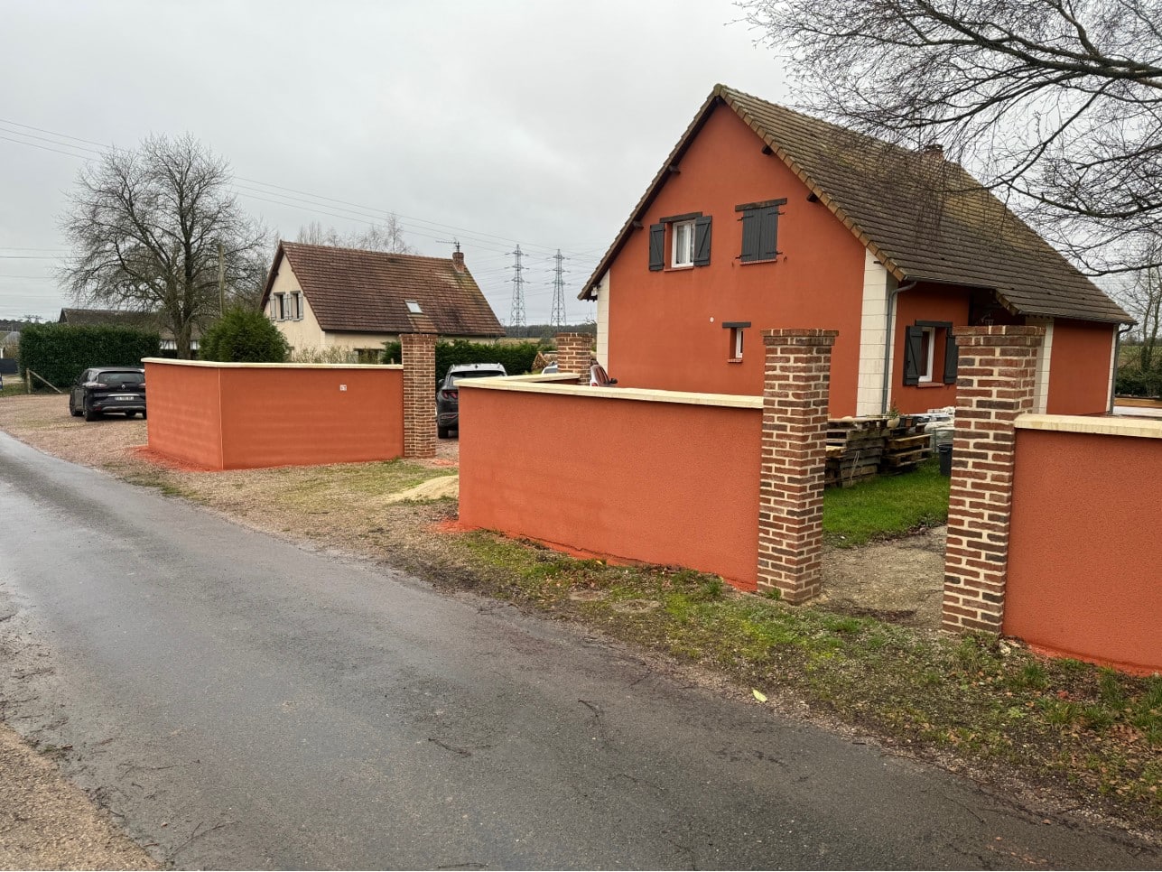 Une maison en brique rouge avec des clôtures en béton rouge entourant la propriété, sous un ciel nuageux.