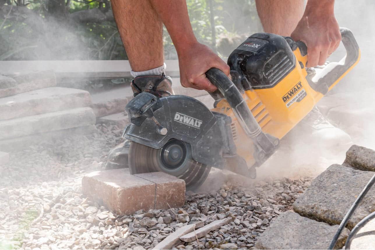 Gros plan sur un ouvrier utilisant une scie circulaire DeWalt pour couper un pavé, générant beaucoup de poussière.