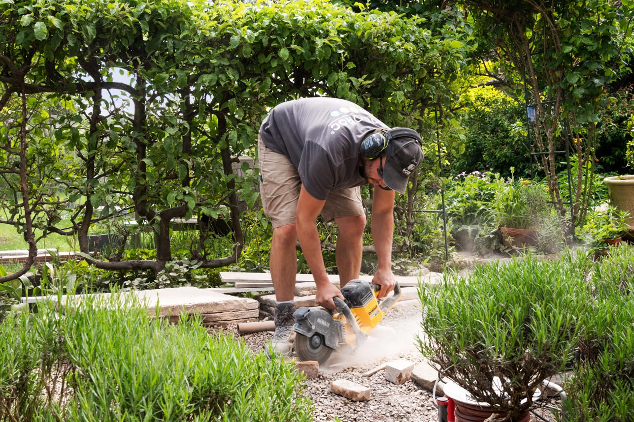 Un ouvrier utilisant une scie circulaire pour couper des pavés dans un jardin, entouré de plantes vertes.